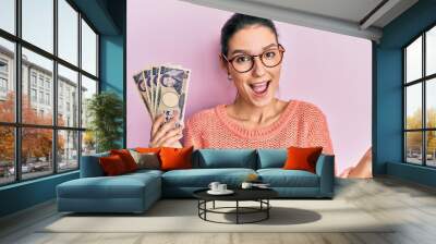 Young caucasian woman holding japanese yen banknotes celebrating achievement with happy smile and winner expression with raised hand Wall mural