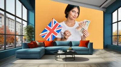 Young caucasian woman holding uk flag and pounds banknotes relaxed with serious expression on face. simple and natural looking at the camera. Wall mural