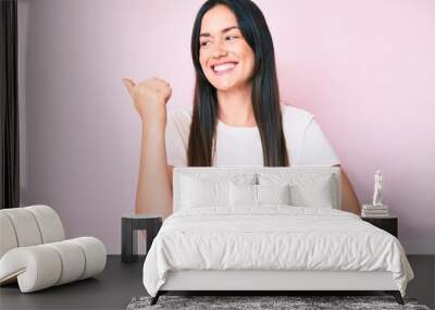 Young caucasian woman sitting at the table wearing casual white tshirt smiling with happy face looking and pointing to the side with thumb up. Wall mural