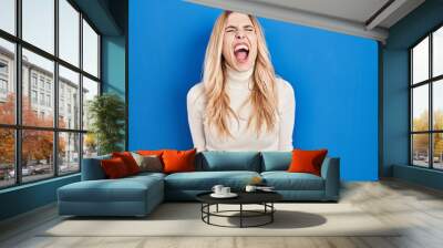 Young caucasian woman standing over blue background angry and mad screaming frustrated and furious, shouting with anger. rage and aggressive concept. Wall mural