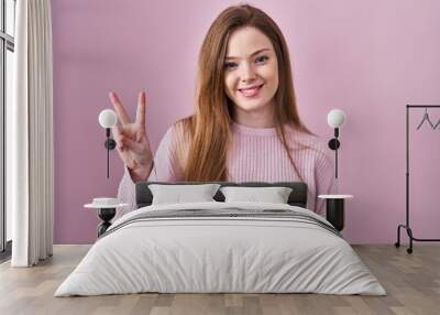 Young caucasian woman standing over pink background showing and pointing up with fingers number two while smiling confident and happy. Wall mural