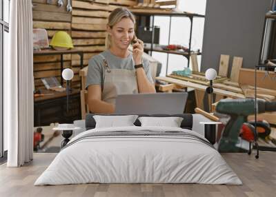 Young caucasian woman talks on phone at a carpentry workshop with laptop and tools. Wall mural