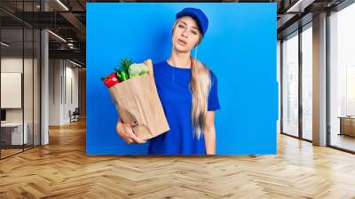 Young caucasian woman wearing courier uniform with groceries from supermarket looking sleepy and tired, exhausted for fatigue and hangover, lazy eyes in the morning. Wall mural