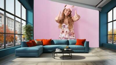 Young caucasian woman wearing sleep mask and pajama approving doing positive gesture with hand, thumbs up smiling and happy for success. winner gesture. Wall mural