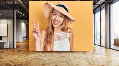 Young caucasian woman wearing summer hat showing and pointing up with fingers number two while smiling confident and happy. Wall mural