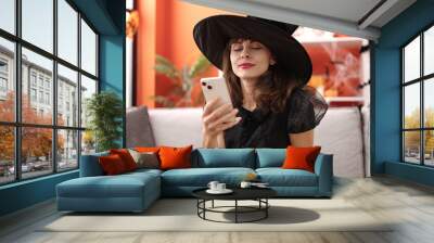 Young caucasian woman wearing witch costume sitting on the sofa with smartphone at home Wall mural