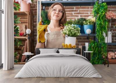 Young caucasian woman working at florist shop holding plant smiling looking to the side and staring away thinking. Wall mural