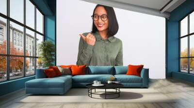 Young chinese woman wearing green shirt and glasses over isolated white background smiling with happy face looking and pointing to the side with thumb up. Wall mural