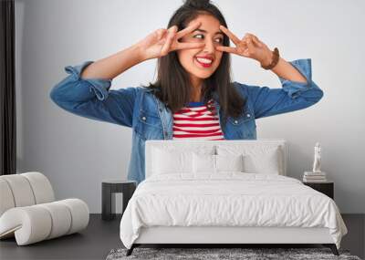 Young chinese woman wearing striped t-shirt and denim shirt over isolated white background Doing peace symbol with fingers over face, smiling cheerful showing victory Wall mural