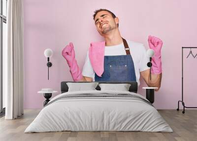 Young cleaner man with blue eyes cleaning wearing apron and gloves over pink background very happy and excited doing winner gesture with arms raised, smiling and screaming for success. Celebration Wall mural