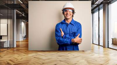 Young constructor man wearing uniform and security helmet over isolated white background happy face smiling with crossed arms looking at the camera. Positive person. Wall mural