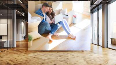 Young couple sitting on the floor of new house around cardboard boxes using laptop, smiling happy for new apartment Wall mural