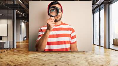 Young detective man looking through magnifying glass over isolated background with a confident expression on smart face thinking serious Wall mural