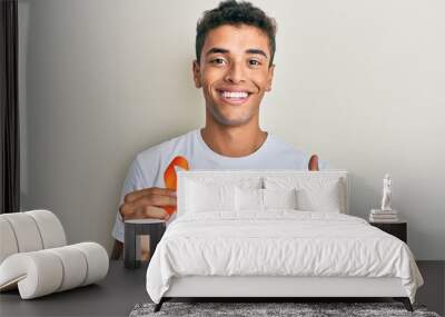 Young handsome african american man holding awareness orange ribbon smiling happy and positive, thumb up doing excellent and approval sign Wall mural