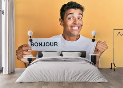 Young handsome african american man holding bonjour french greeting word screaming proud, celebrating victory and success very excited with raised arm Wall mural