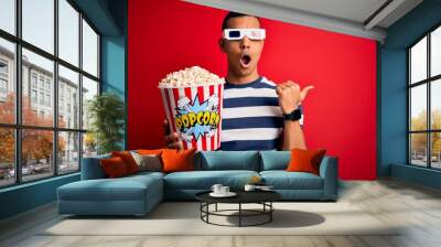 Young handsome african american man watching movie using 3d glasses eating popcorns Surprised pointing with hand finger to the side, open mouth amazed expression. Wall mural