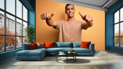 Young handsome african american man wearing casual t-shirt standing over yellow background approving doing positive gesture with hand, thumbs up smiling and happy for success. Winner gesture. Wall mural