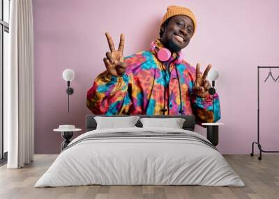 Young handsome african american man wearing colorful coat and cap over pink background smiling looking to the camera showing fingers doing victory sign. Number two. Wall mural