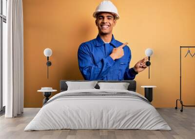 Young handsome african american worker man wearing blue uniform and security helmet smiling and looking at the camera pointing with two hands and fingers to the side. Wall mural