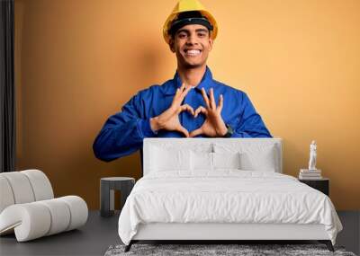 Young handsome african american worker man wearing blue uniform and security helmet smiling in love doing heart symbol shape with hands. Romantic concept. Wall mural