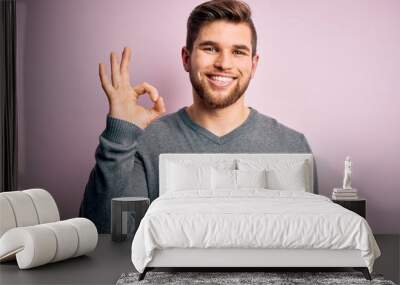 Young handsome blond man with beard and blue eyes wearing casual sweater smiling positive doing ok sign with hand and fingers. Successful expression. Wall mural