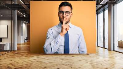 Young handsome businessman wearing tie and glasses standing over yellow background asking to be quiet with finger on lips. Silence and secret concept. Wall mural