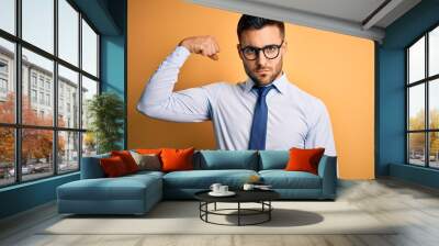 Young handsome businessman wearing tie and glasses standing over yellow background Strong person showing arm muscle, confident and proud of power Wall mural