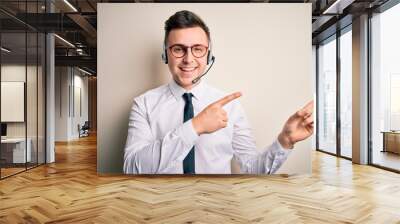 Young handsome caucasian business man wearing call center headset at customer service smiling and looking at the camera pointing with two hands and fingers to the side. Wall mural