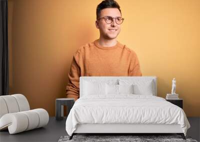 Young handsome caucasian man wearing glasses and casual winter sweater over yellow background looking away to side with smile on face, natural expression. Laughing confident. Wall mural