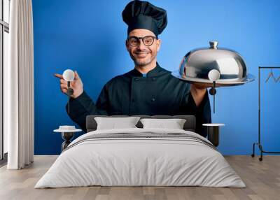 Young handsome chef man wearing uniform and hat holding waiter tray with dome very happy pointing with hand and finger to the side Wall mural