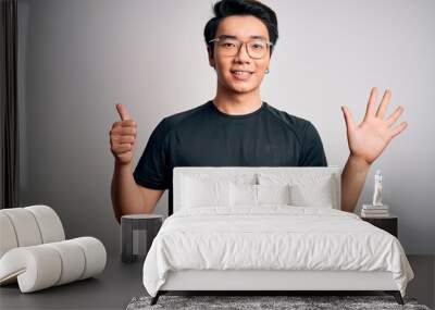 Young handsome chinese man wearing black t-shirt and glasses over white background showing and pointing up with fingers number six while smiling confident and happy. Wall mural