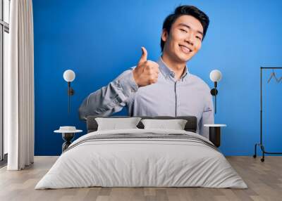 Young handsome chinese man wearing casual shirt standing over isolated blue background doing happy thumbs up gesture with hand. Approving expression looking at the camera showing success. Wall mural