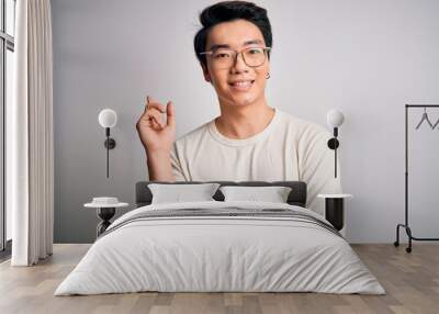 Young handsome chinese man wearing casual t-shirt and glasses over white background with a big smile on face, pointing with hand and finger to the side looking at the camera. Wall mural