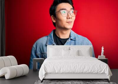 Young handsome chinese man wearing denim jacket and glasses over red background smiling looking to the side and staring away thinking. Wall mural