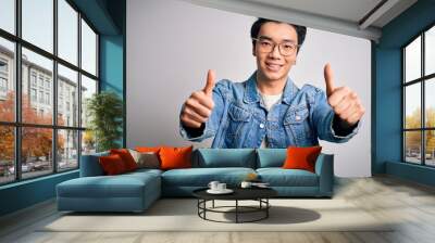 Young handsome chinese man wearing denim jacket and glasses over white background approving doing positive gesture with hand, thumbs up smiling and happy for success. Winner gesture. Wall mural