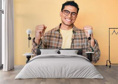 Young handsome hispanic man wearing casual clothes and glasses screaming proud, celebrating victory and success very excited with raised arms Wall mural