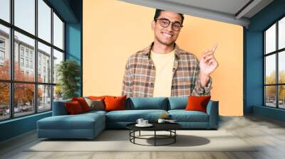 Young handsome hispanic man wearing casual clothes and glasses smiling happy pointing with hand and finger to the side Wall mural