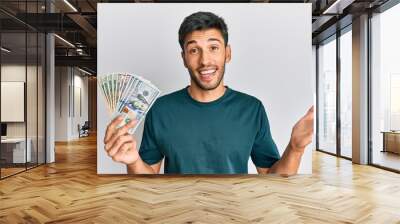 Young handsome man holding dollars celebrating achievement with happy smile and winner expression with raised hand Wall mural