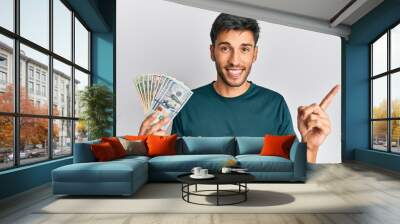 Young handsome man holding dollars smiling happy pointing with hand and finger to the side Wall mural