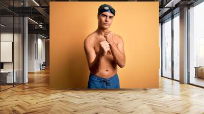 Young handsome man shirtless wearing swimsuit and swim cap over isolated yellow background Ready to fight with fist defense gesture, angry and upset face, afraid of problem Wall mural