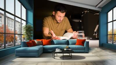 Young handsome man studying at home, reading a book at night Wall mural