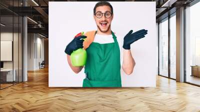 Young handsome man wearing apron holding sprayer celebrating victory with happy smile and winner expression with raised hands Wall mural