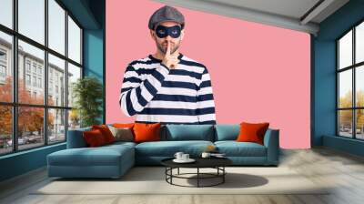 Young handsome man wearing burglar mask asking to be quiet with finger on lips. silence and secret concept. Wall mural