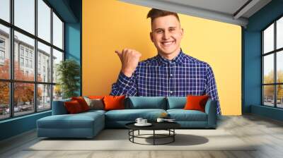 Young handsome man wearing casual shirt standing over isolated yellow background smiling with happy face looking and pointing to the side with thumb up. Wall mural