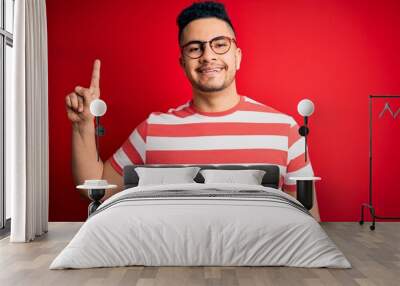 Young handsome man wearing casual striped t-shirt and glasses over isolated red background showing and pointing up with finger number one while smiling confident and happy. Wall mural