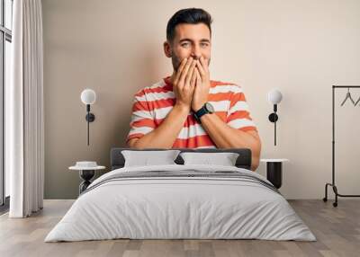 Young handsome man wearing casual striped t-shirt standing over isolated white background laughing and embarrassed giggle covering mouth with hands, gossip and scandal concept Wall mural