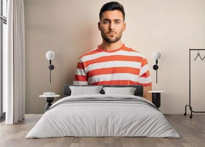 Young handsome man wearing casual striped t-shirt standing over isolated white background Relaxed with serious expression on face. Simple and natural looking at the camera. Wall mural