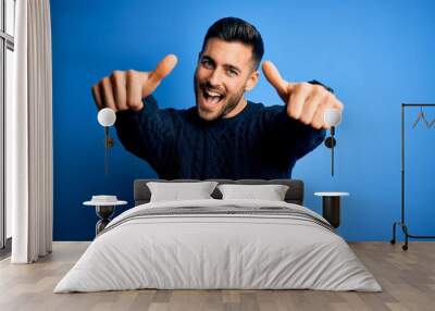 Young handsome man wearing casual sweater standing over isolated blue background approving doing positive gesture with hand, thumbs up smiling and happy for success. Winner gesture. Wall mural