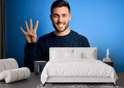 Young handsome man wearing casual sweater standing over isolated blue background showing and pointing up with fingers number four while smiling confident and happy. Wall mural