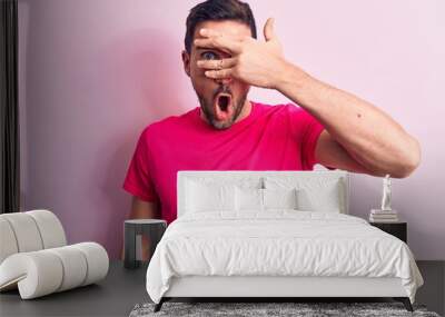 Young handsome man wearing casual t-shirt standing over isolated pink background peeking in shock covering face and eyes with hand, looking through fingers afraid Wall mural